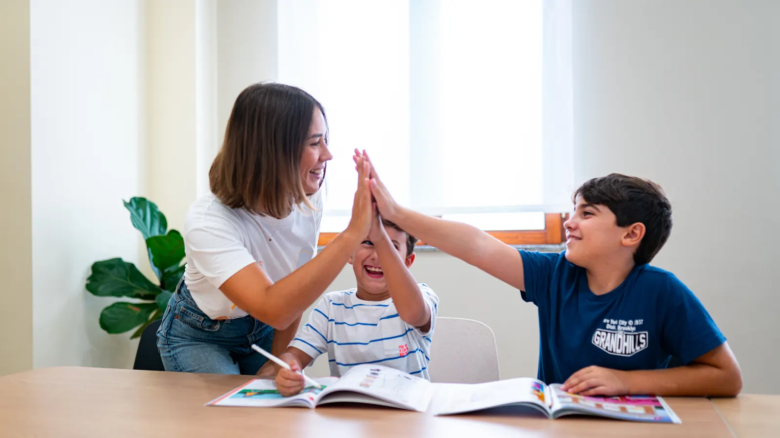 Una teacher batte il cinque a due bambini seduti al tavolo con il quaderno aperto, in un momento di festa in classe