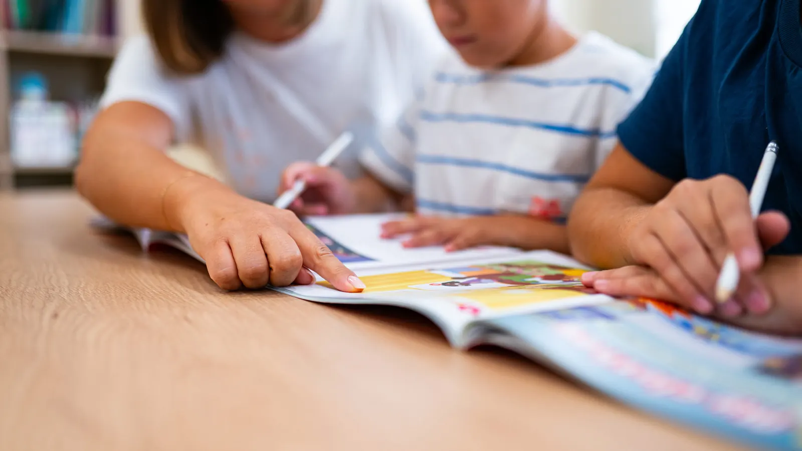 Mani di una teacher e di due bambini che indicano e scrivono su un quaderno di esercizi colorato, sul tavolo di legno