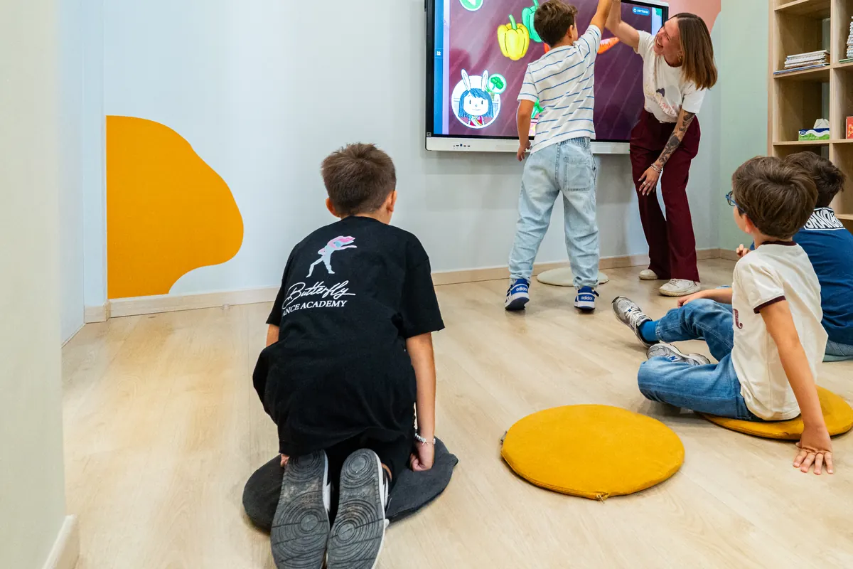 Bambini seduti sui cuscini davanti alla lavagna interattiva, una teacher batte il cinque a un bambino