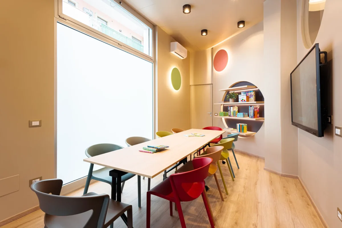 Aula della sede Kokoland di Monopoli con grande tavolo, sedie colorate, libreria rotonda e lavagna interattiva